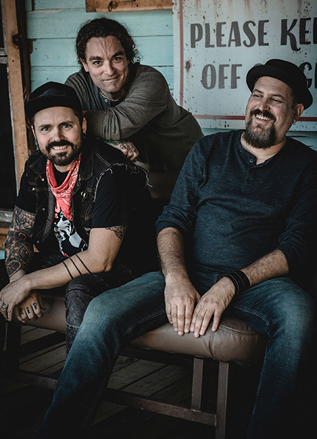 3-member band "The Killbillies" in relaxed poses, wearing affable expressions, seated in from of a wood-panel building.