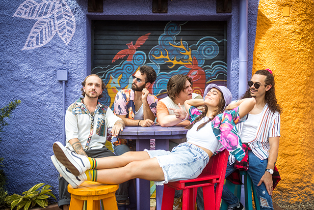 5-member band "Actual Bank Robbers" in relaxed, seated poses in front of colorful muralled wall.