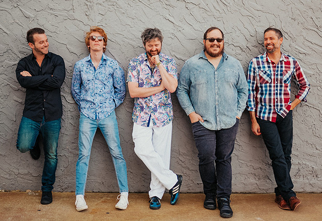 Five members of the band Hot Pink posed side by side against a wall, laughing or grinning.