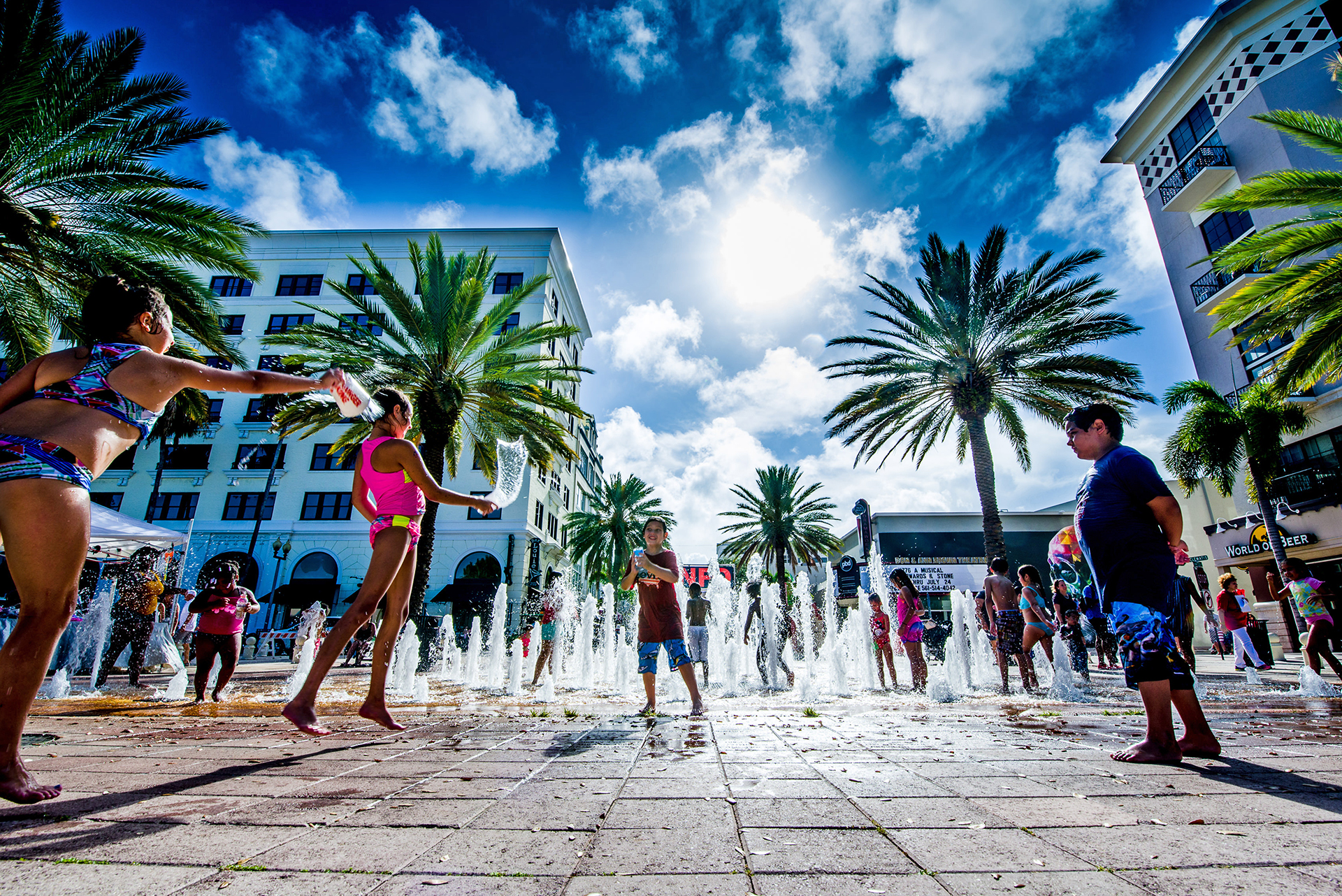 Low angle photo of children playing in fountain under a bright blue sky in the summer, in downtown West Palm Beach's Centennial Square.