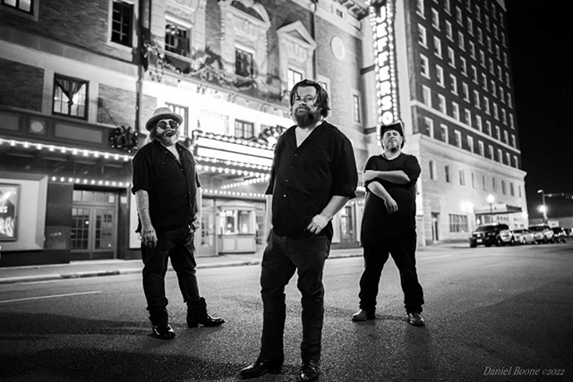 Black and white image of Damon Fowler band members (3 men) in dark clothes, standing on a street in front of a brightly lit downtown theatrer.