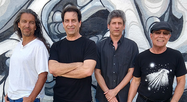 Four male members of Sacred Union band standing side-by-side against a black and white muraled wall.