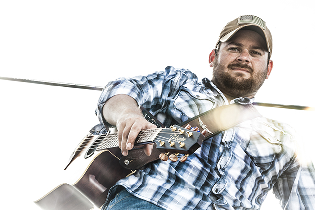 Up-shot of bearded, ball-capped musician, Andrew Morris, holding a guitar, looking directly at viewer with neutral expression.