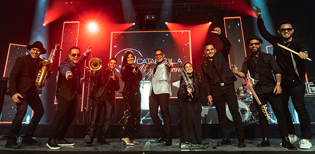 Dramatically lit, fish-eye image of exuberant 9-member Catabella Band on stage, posing with smiles, instruments and arms aloft.