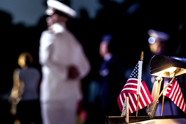 Navy officer in dress whites standing at attention in the distance, an American flag in sharp focus in foreground.