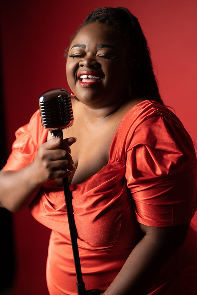 Portrait of CeCe Teneal in elegant red dress on red background, singing into a vintage microphone