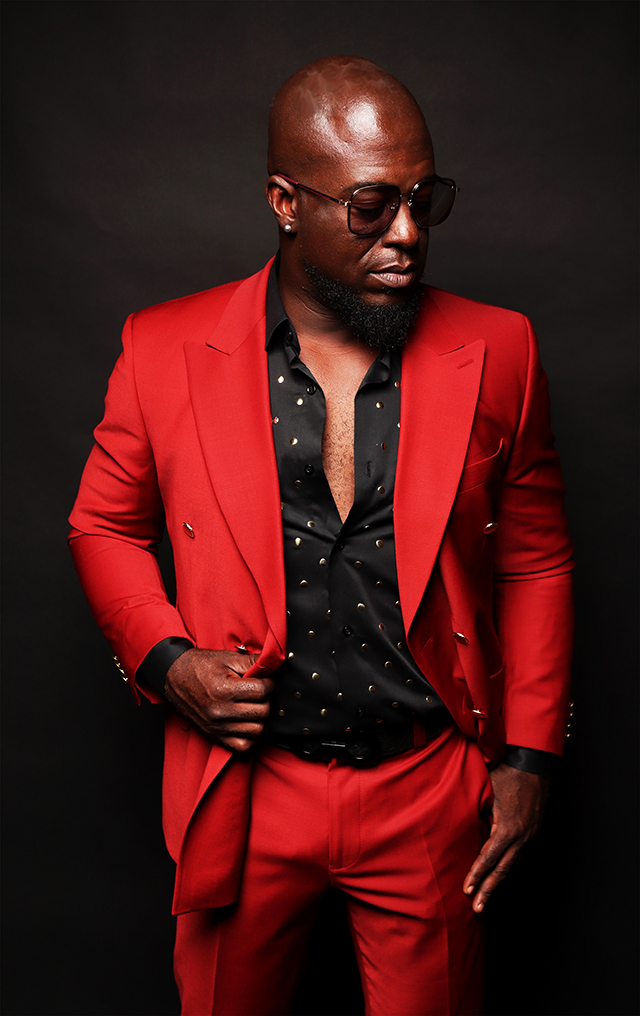 Dramatic portrait of Jazz musician Nathan Mitchell, posed wearing sunglasses, in red suit, against a dark background.