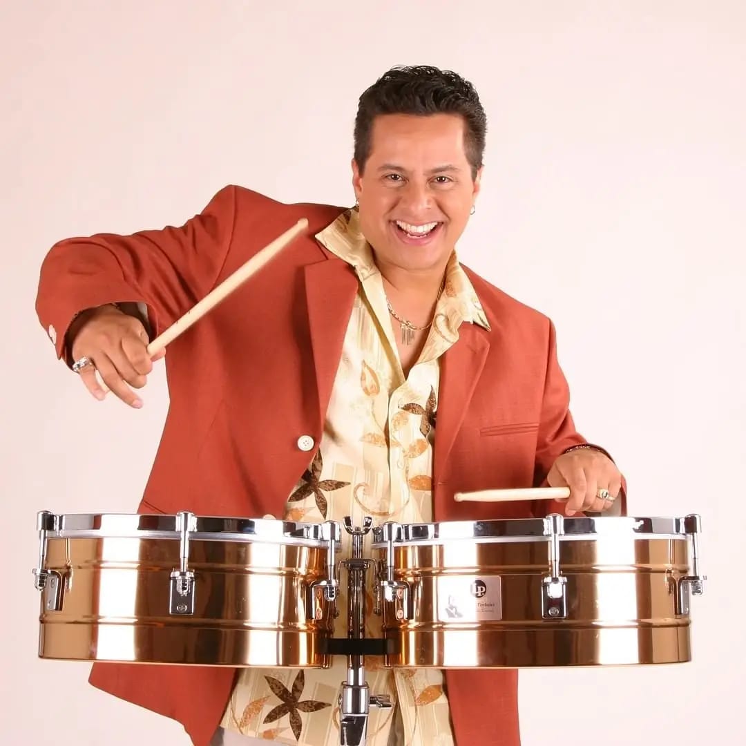 Studio photo of Tito Puente Jr. smiling enthusiastically while playing the drums