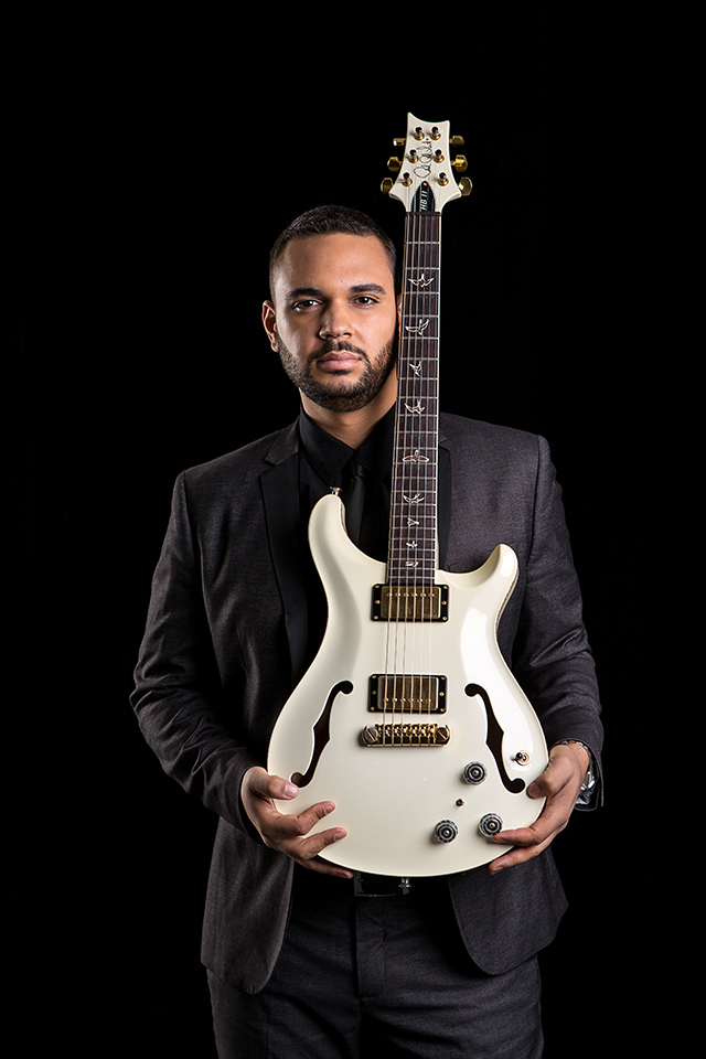 Elegant portrait of Jazz musician Adam Hawley holding a white guitar at his waist, with neck resting next to his face and head. He wears a suit, and is standing on a deep black background.