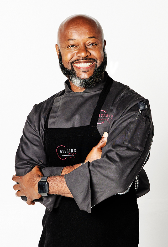 Portrait of Chef Winston Williams with a frank, engaging smile, posed with arms crossed in black apron and shirt against a white background.