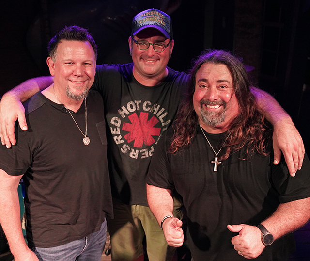 3 members of The Happiness Club band, smiling, posed against black background.