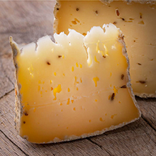 Close-up photo of a fresh cheese wedge cut from a round behind it, displayed on a wooden cutting board.