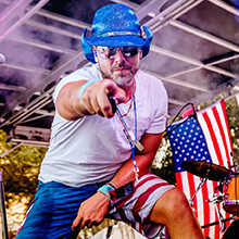 Photo of musical performer on stage, wearing blue cowboy hat with flags behind him, pointing directly at viewer