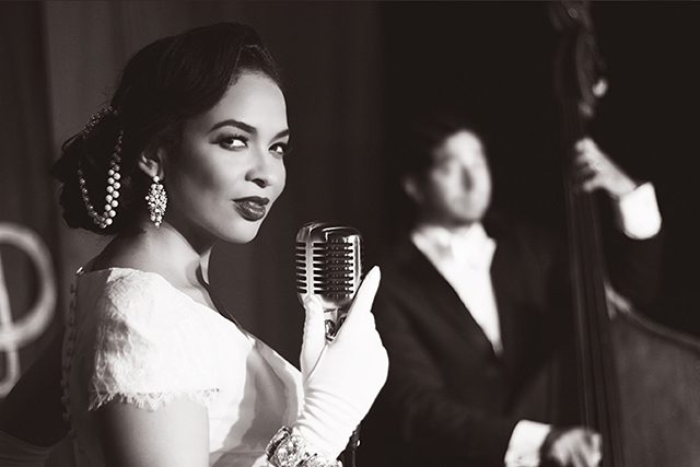 Black and white image of Jazz vocalist Oya in a sultry pose singing into a vintage microphone on a 1940's-styled nightclub stage